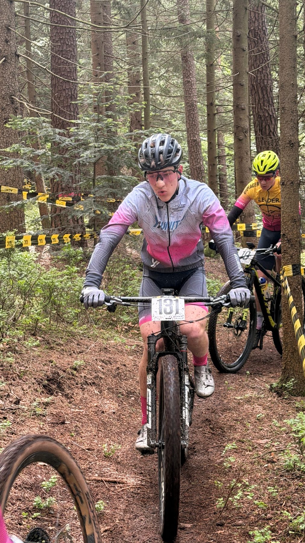 Fahrer mit der Startnummer 131 kämpft sich bei Nässe durch einen Nadelwald, dahinter eine Fahrerin im Leadertrikot.